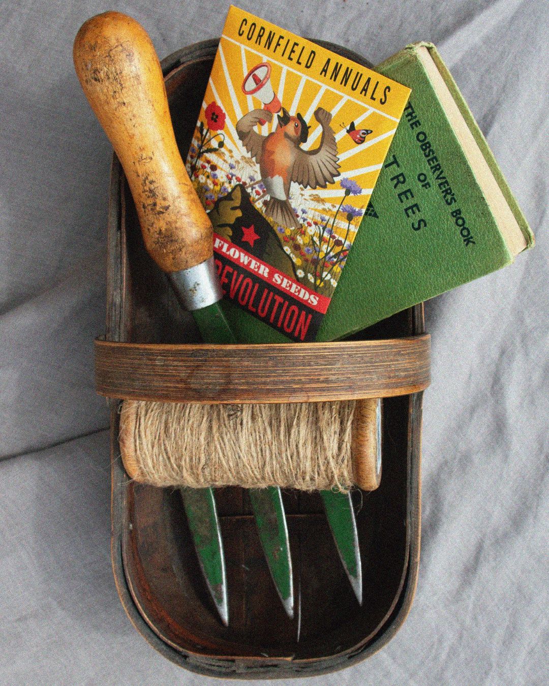 Set of gardening tools in a wooden caddy with a cornfield annuals wildflower seed packet and book on a grey background. Showcasing Seed Revolution's Cornfield Annuals Wildflower Seed Mix, british wild flower seeds, british wildflower seeds, wildflower mixes, mixed wildflower seeds, uk wildflower seeds, wildflower meadows, wildflower meadow, annual wildflowers, Cornfield Annuals, cornfield uk.
