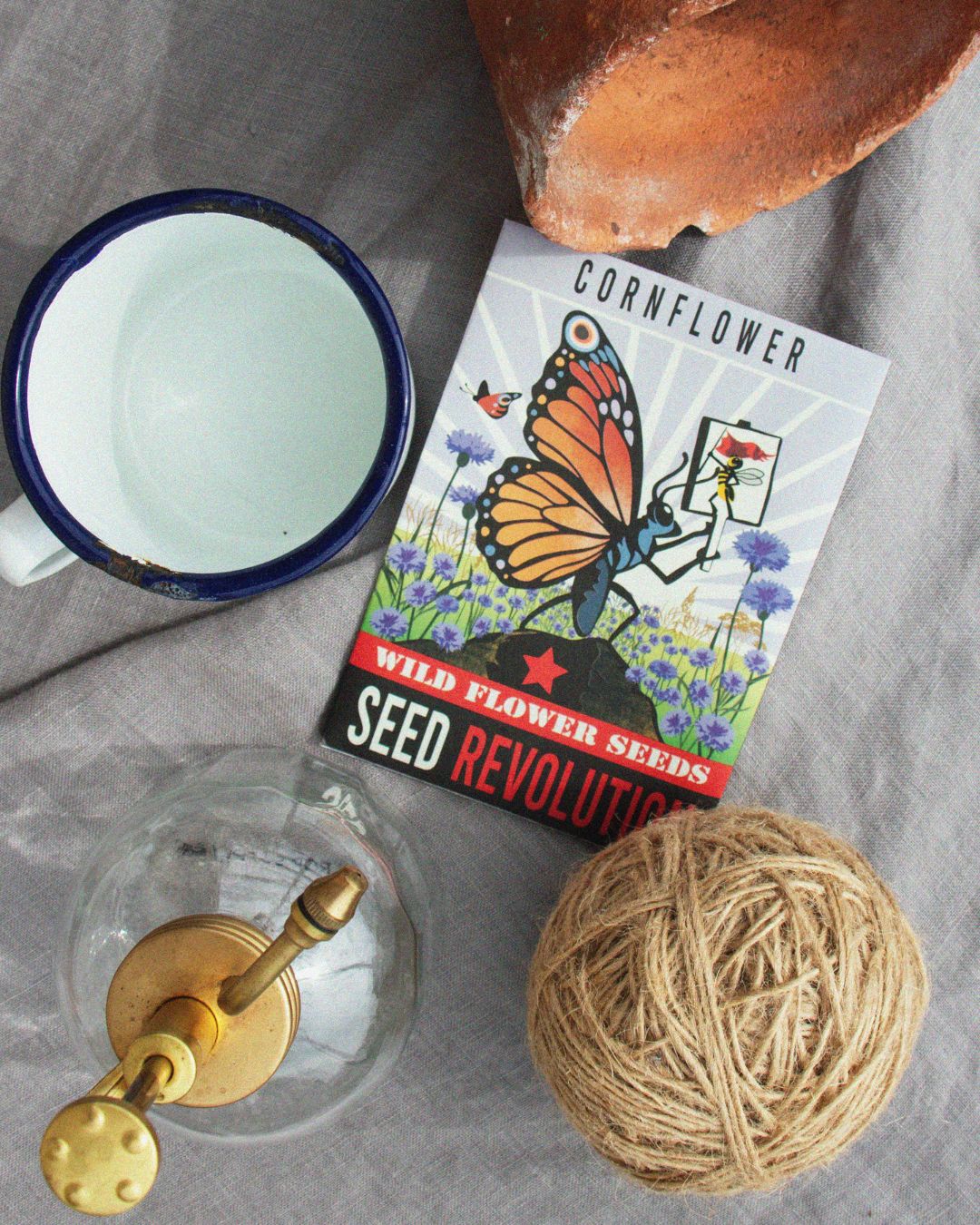 A cornflower wildflower seed packet of surrounded by a gardening items including a terracotta pot on a grey fabric background. Showcasing Seed Revolution's Cornflower, Centaurea cyanus, cornflowers, corn flowers, cornflower blue, british wild flower seeds, british wildflower seeds, uk wildflower seeds.