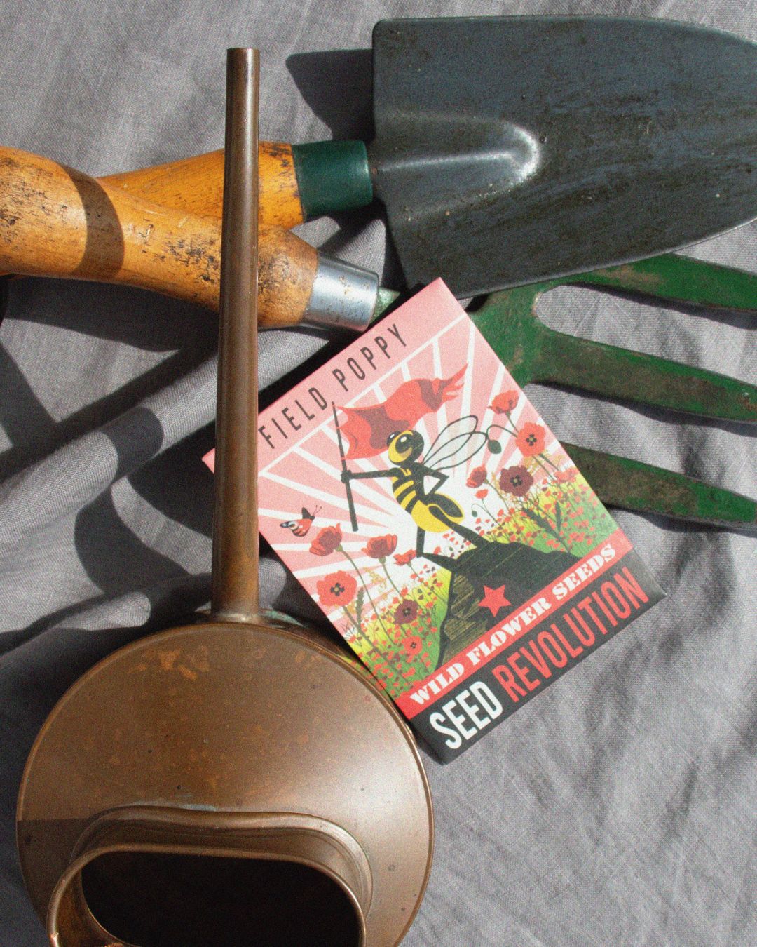A poppy wildflower seed packet labeled lies on grey fabric, surrounded by gardening tools. Showcasing Seed Revolution's Field Poppy, Papaver rhoeas, poppy flower, poppy flowers, wildflower campion, british wild flower seeds, british wildflower seeds, uk wildflower seeds.