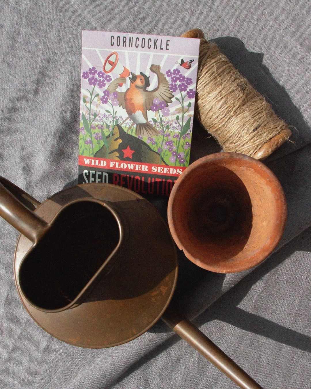 Corncockle wildflower seed packet with gardening items on a grey surface. Showcasing Seed Revolution's Corncockle, Agrostemma githago, corn cockle, Agrostemma cockle, british wild flower seeds, british wildflower seeds, uk wildflower seeds.