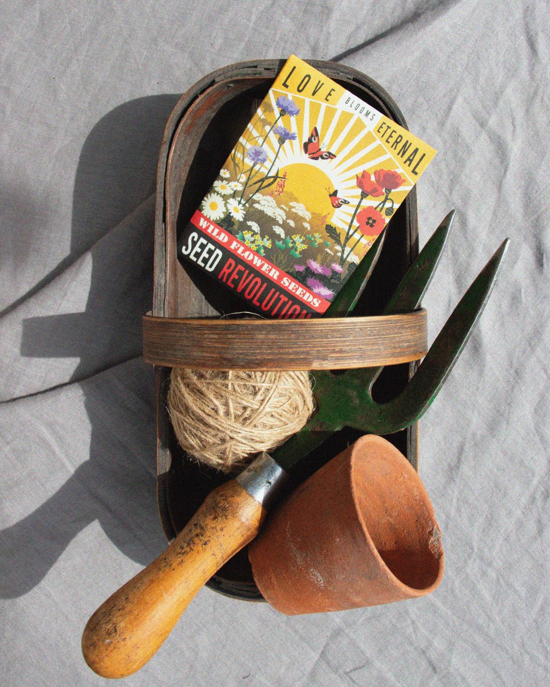 Gardening tools and a Love Blooms Eternal wildflower mix seed starter card on a grey fabric background. Showcasing Seed Revolution's Native Plant Pot Wildflower Seed Mix, british wild flower seeds, british wildflower seeds, wildflower mixes, mixed wildflower seeds, uk wildflower seeds, wildflower meadows, wildflower meadow, universal wildflowers, wildflowers for pots, cost-effective wildflower meadow.