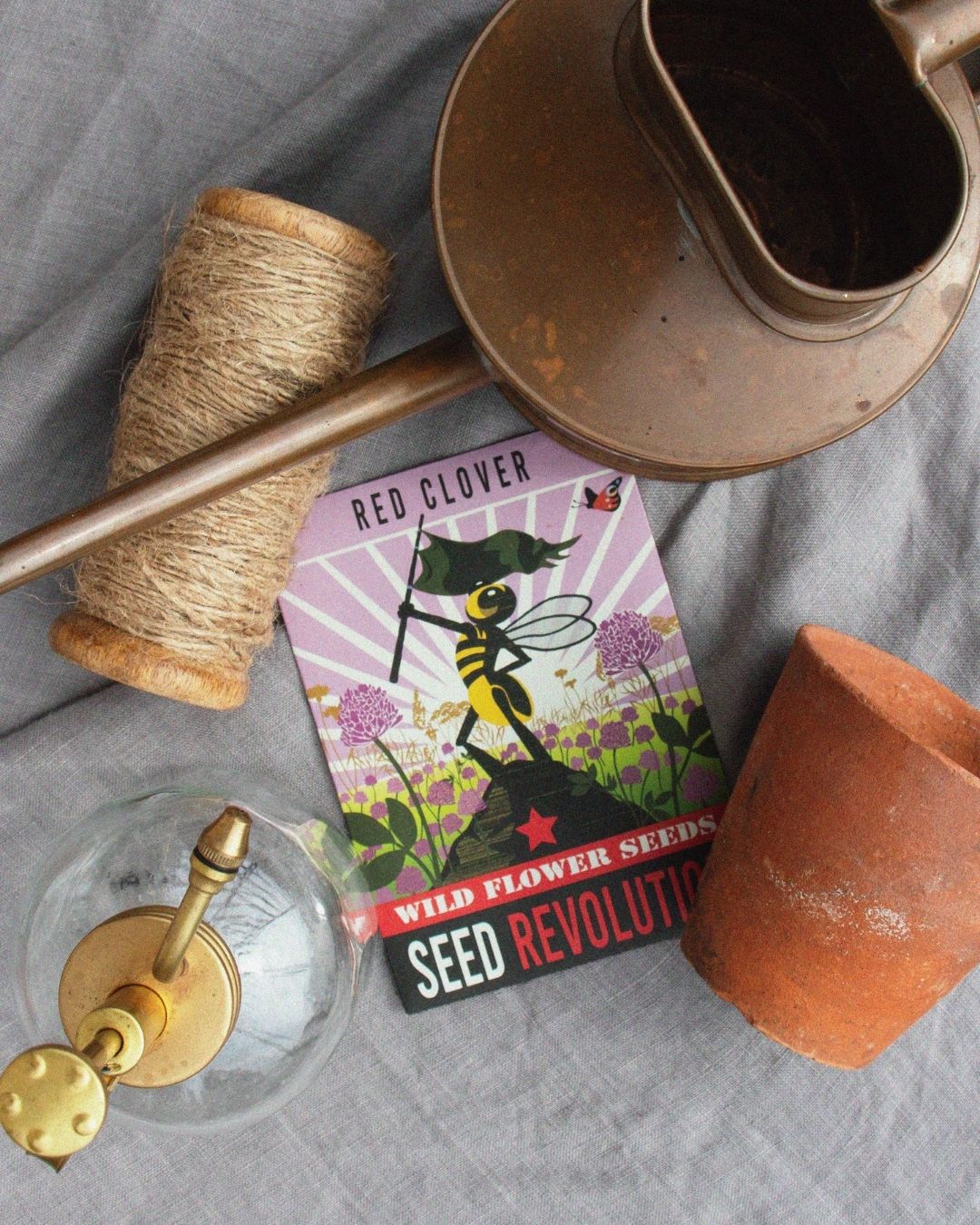 Wildflower Red Clover Seed packet with a bee illustration, surrounded by rustic gardening items on a grey surface. Showcasing Seed Revolution's Trifolium pratense, clover red, red clover Trifolium, british wild flower seeds, british wildflower seeds, uk wildflower seeds.